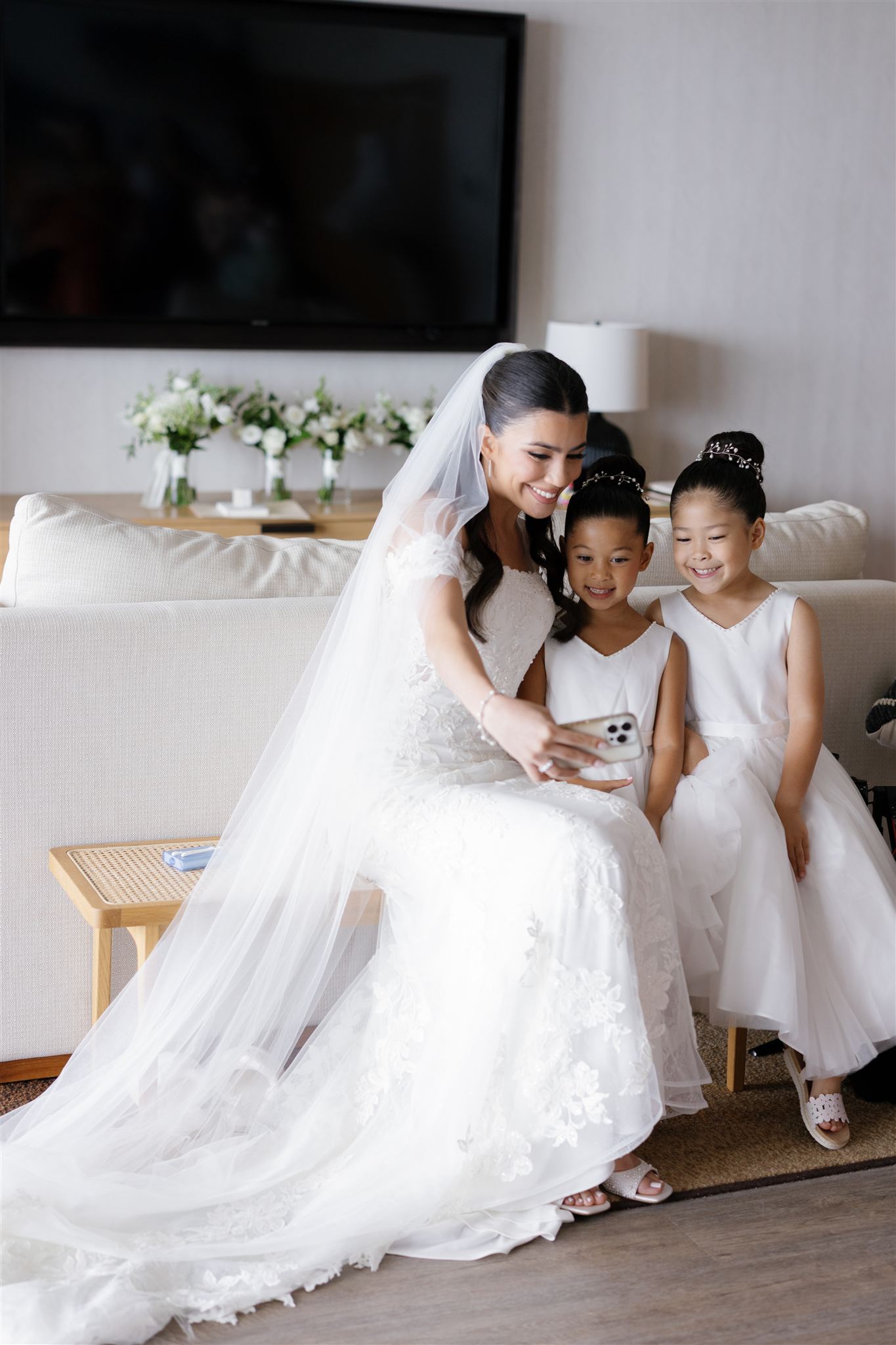 a bride sitting on a wooden bench smiling and taking a selfie with her two flower girls 