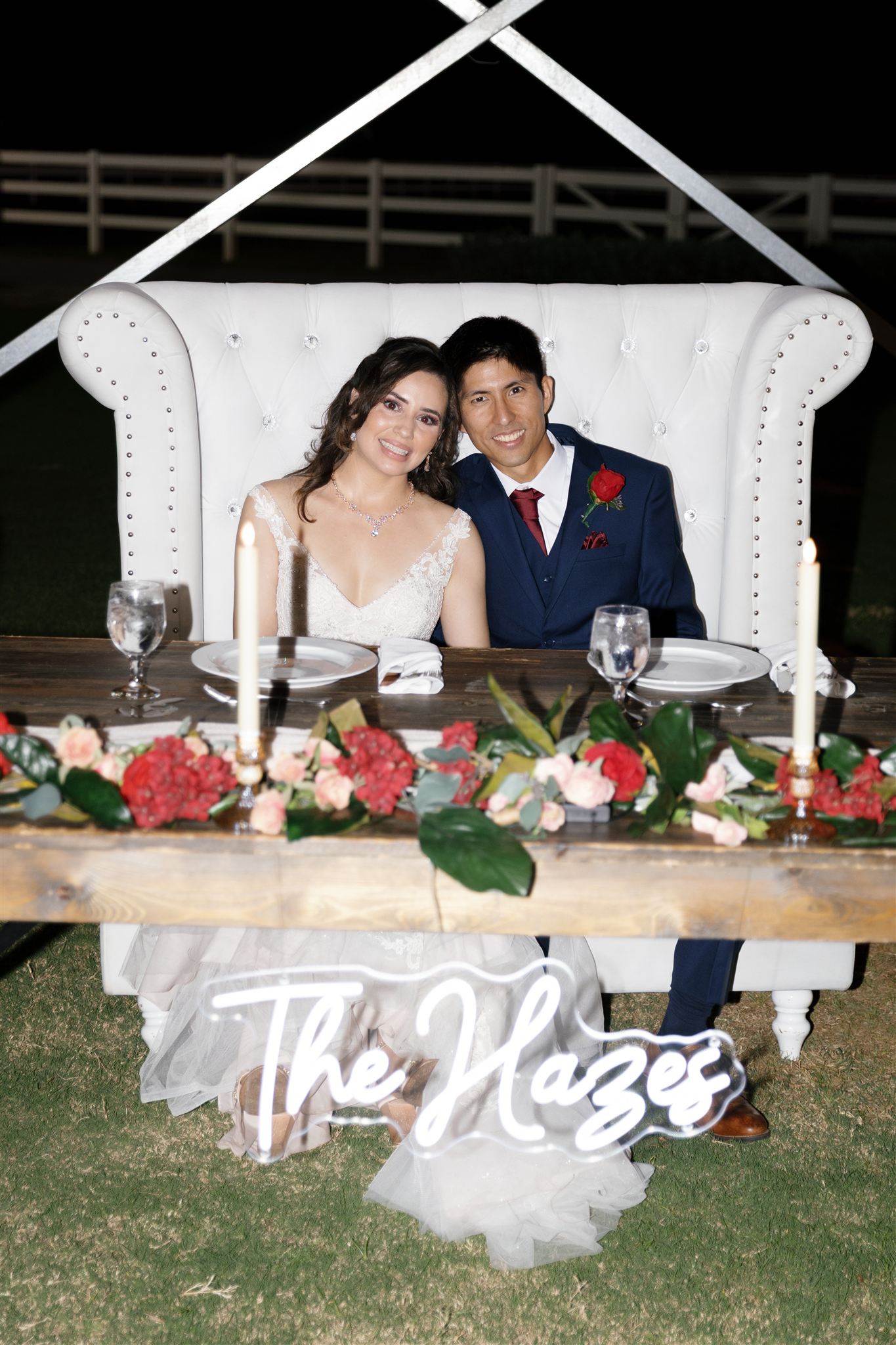 a couple sitting next to each other their wedding reception in a white chair in front of them is a long wooden table decorated with flowers, plates, and candles 