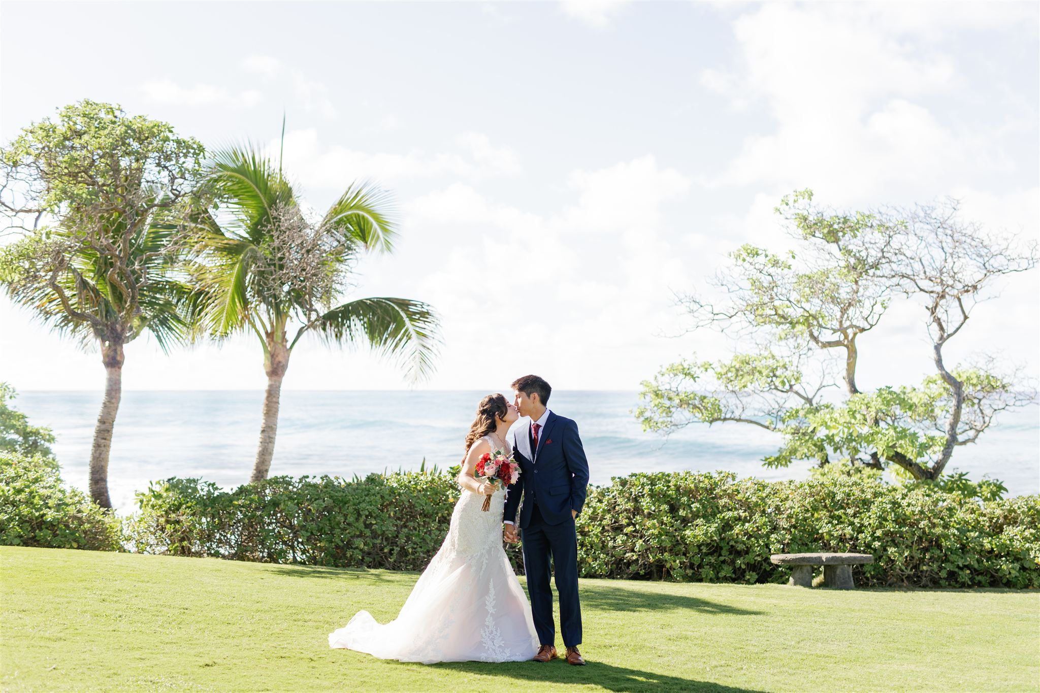 a couple standing on the lawn of Ritz-Carlton O'ahu, Turtle Bay next to the ocean kissing one another on the day of their wedding 