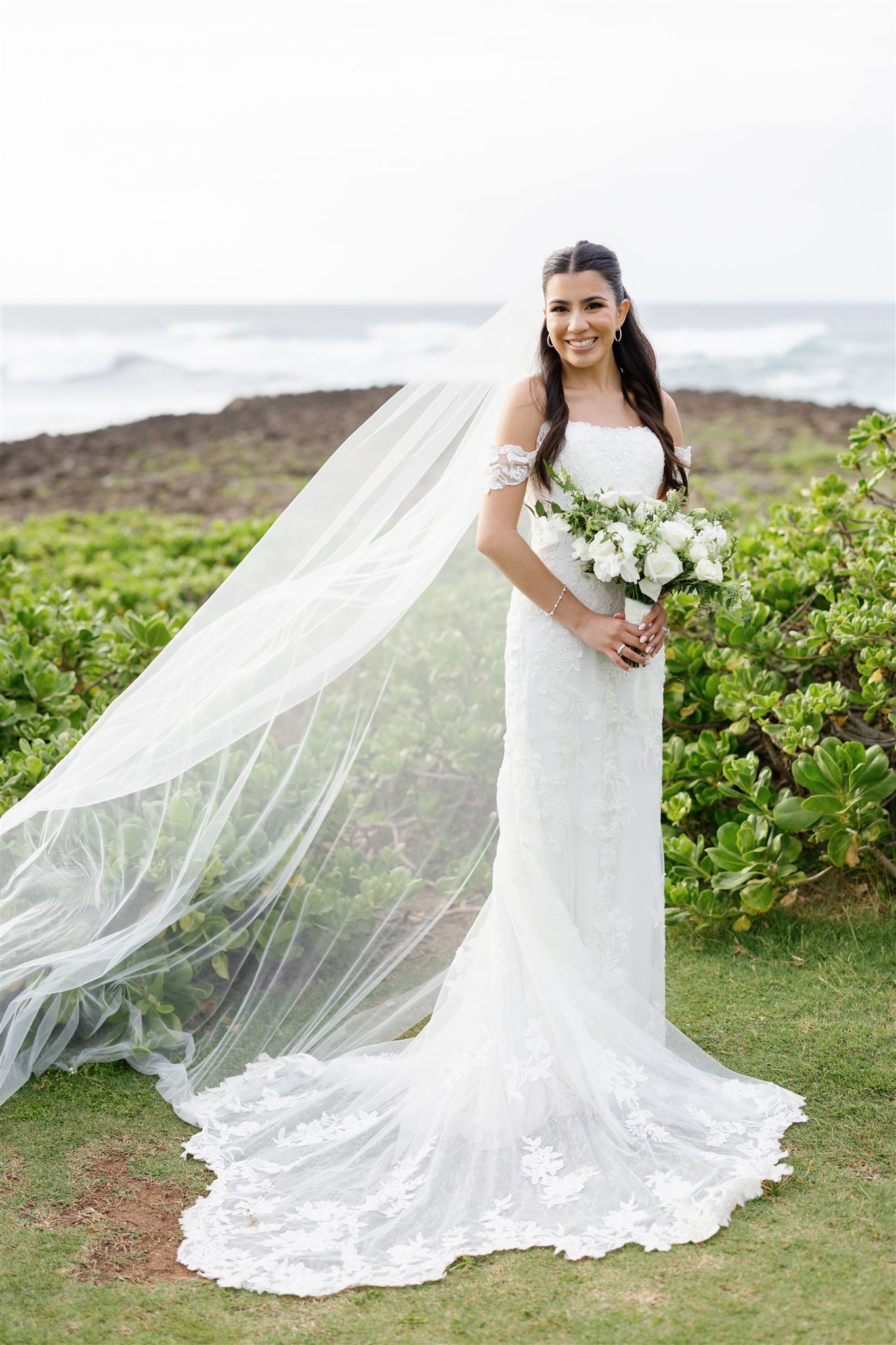 a bride standing next to the ocean and smiling and holding a wedding bouquet 