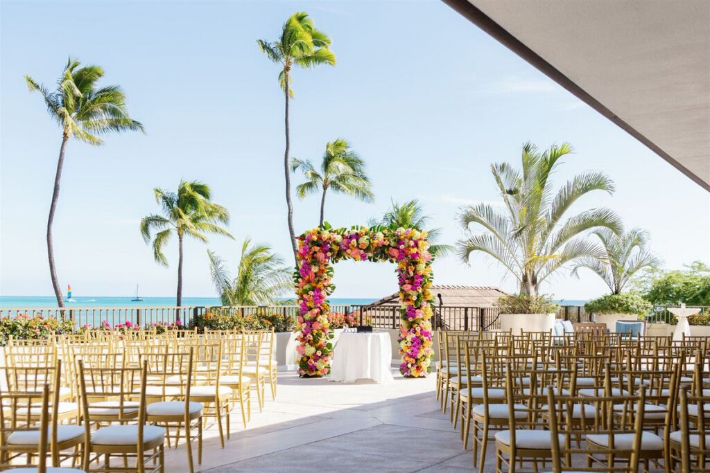 A colorful floral arch at the end of a small aisle on a terrace set up for a wedding ceremony 