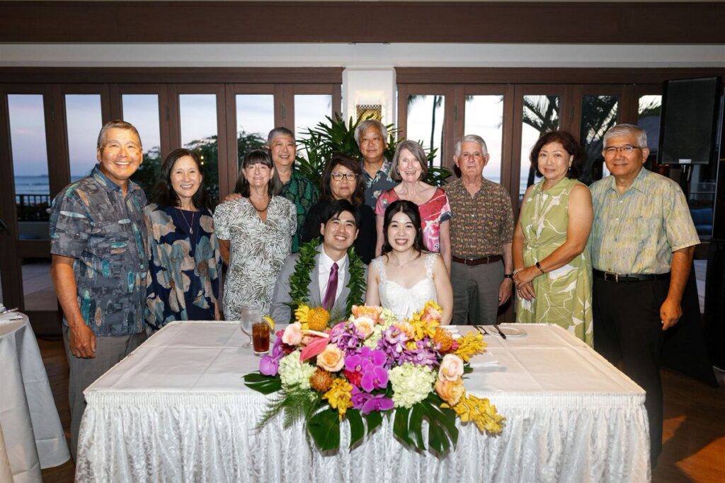 Newlyweds sitting at a table with a colorful floral arrangement with their families smiling behind them 
