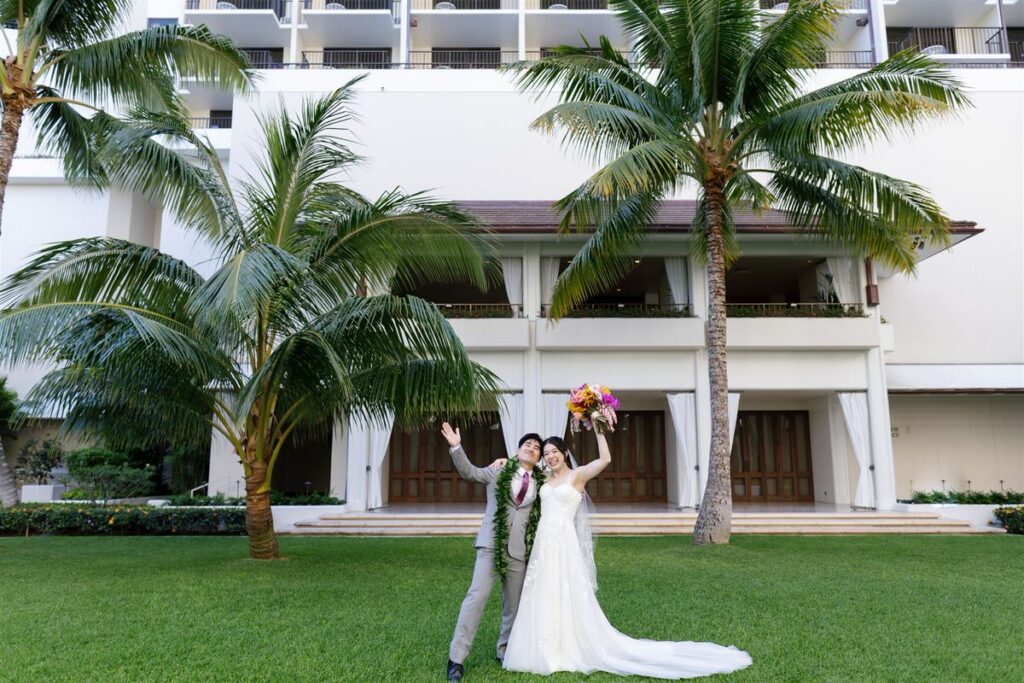 A newlywed couple standing in front of a white hotel with palm trees out from with their arms outstretched 