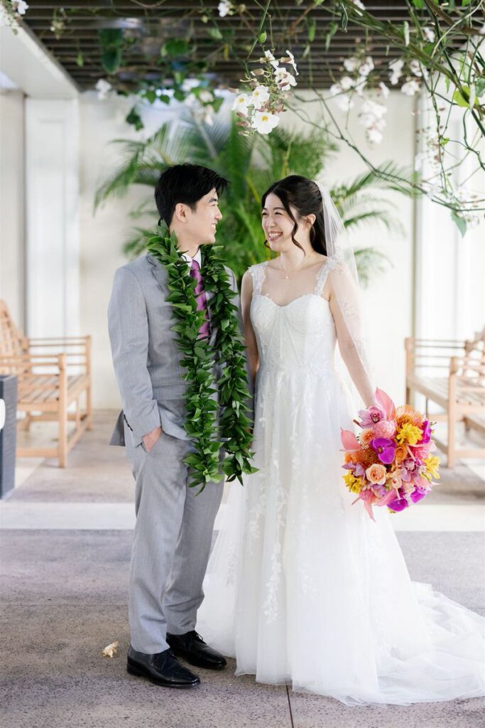 Newlyweds smiling at each other as one wears a long green lei and the other holds a colorful bouquet of flowers 