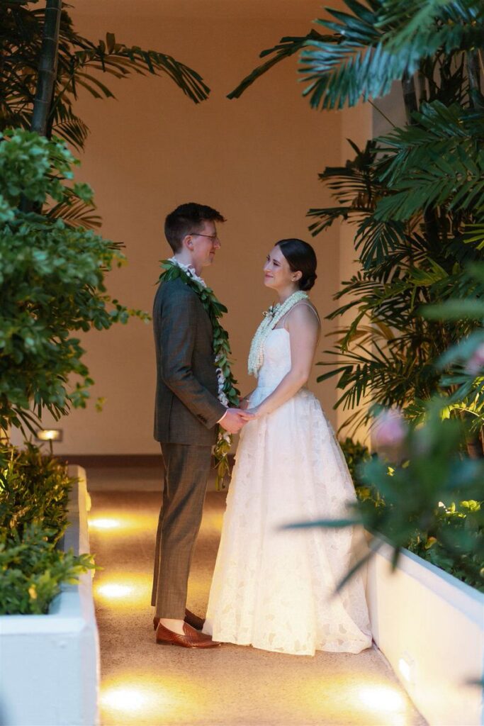 Newlyweds holding hands in a dimly lit hallway while smiling at each other 