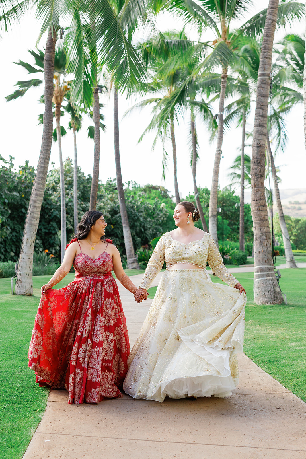 two women walking hand in hand down a paved path underneath the palm trees smiling at each other 
