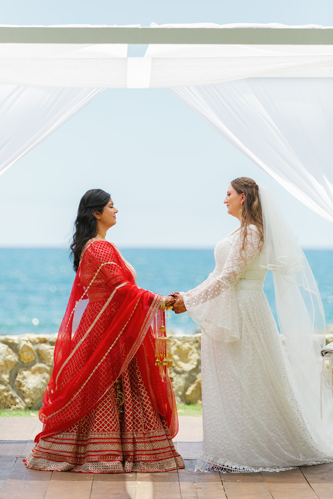 two women having a wedding ceremony holding hands and smiling at each other the ceremony is taking place next to the ocean