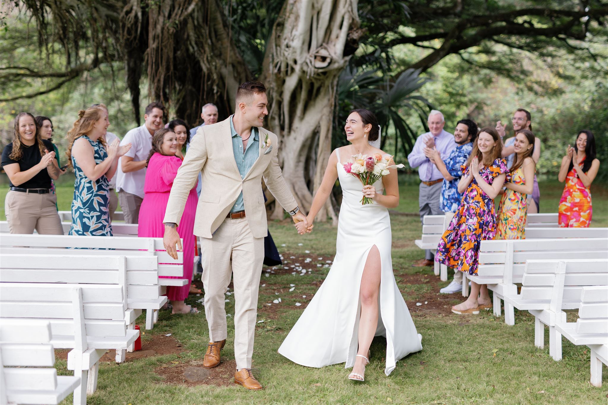 a bride and groom holding hands and laughing with each other after their wedding ceremony at palm meadow in waimea valley as their guests cheer for them