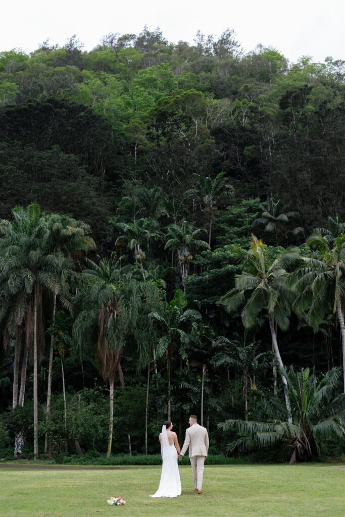 a bride and groom holding hands and walking towards the palm trees in waimea valley
