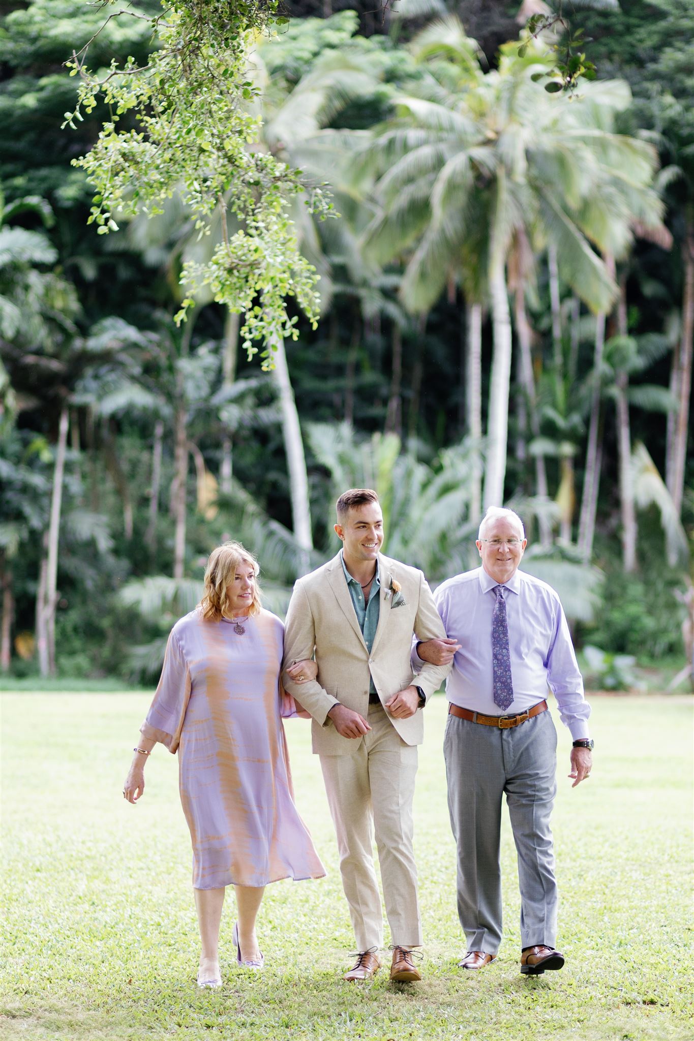 a groom walking down the lawn at palm meadow in waimea valley arm in arm with his parents