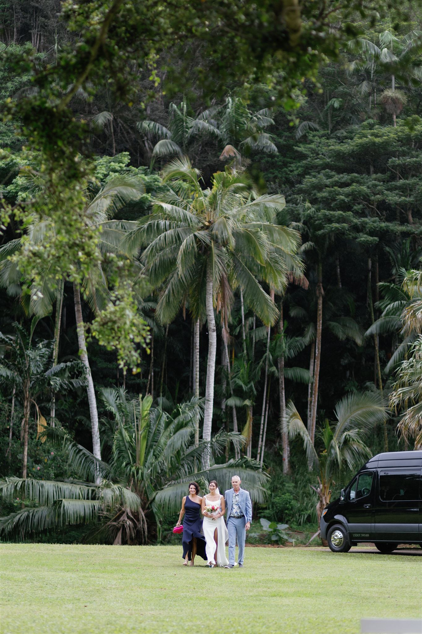 a bride walking down the lawn at palm meadow in waimea valley with her mother and father next to her