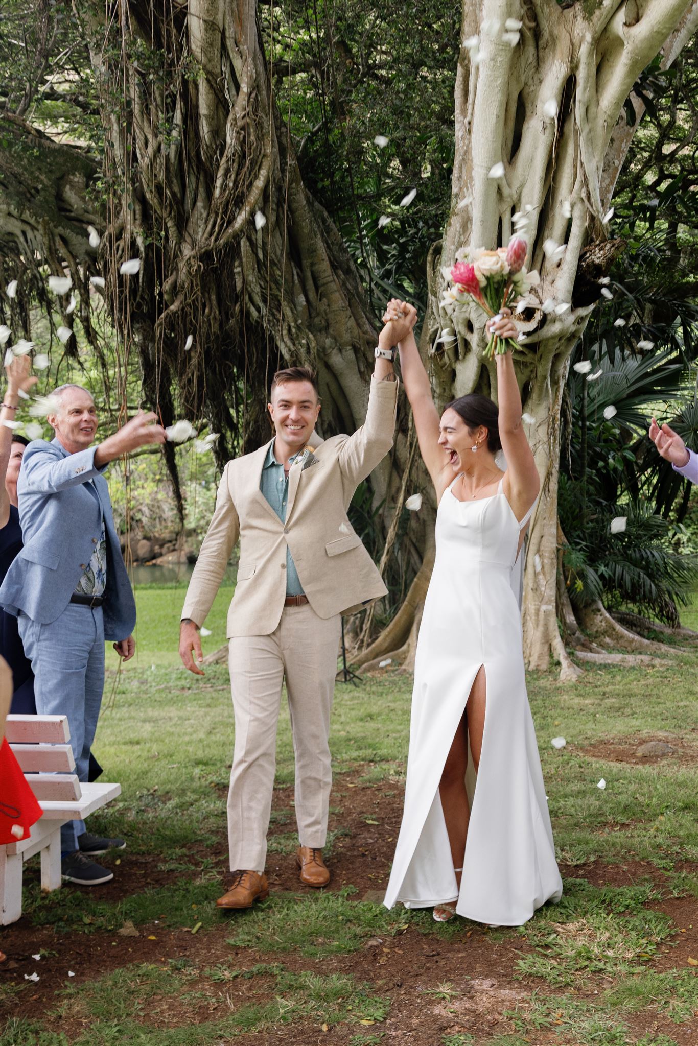 a bride and groom celebrating at their wedding ceremony at waimea valley as their guests throw petals above their head