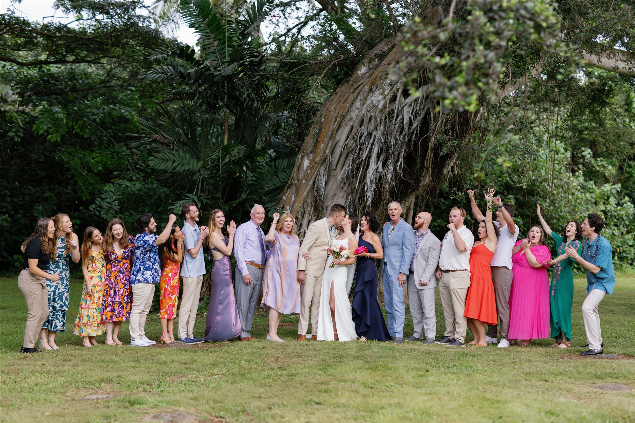 a bride and groom kissing in palm meadow in waimea valley while their wedding guests stand next to them and cheer for them