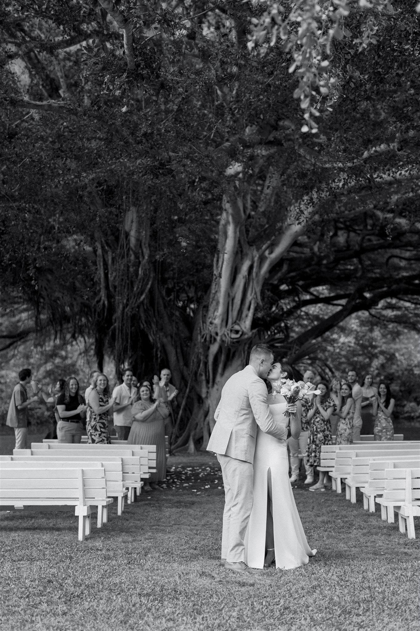 a bride and groom kissing and embracing in front of a large tree in waimea valley as their wedding guests cheer for them