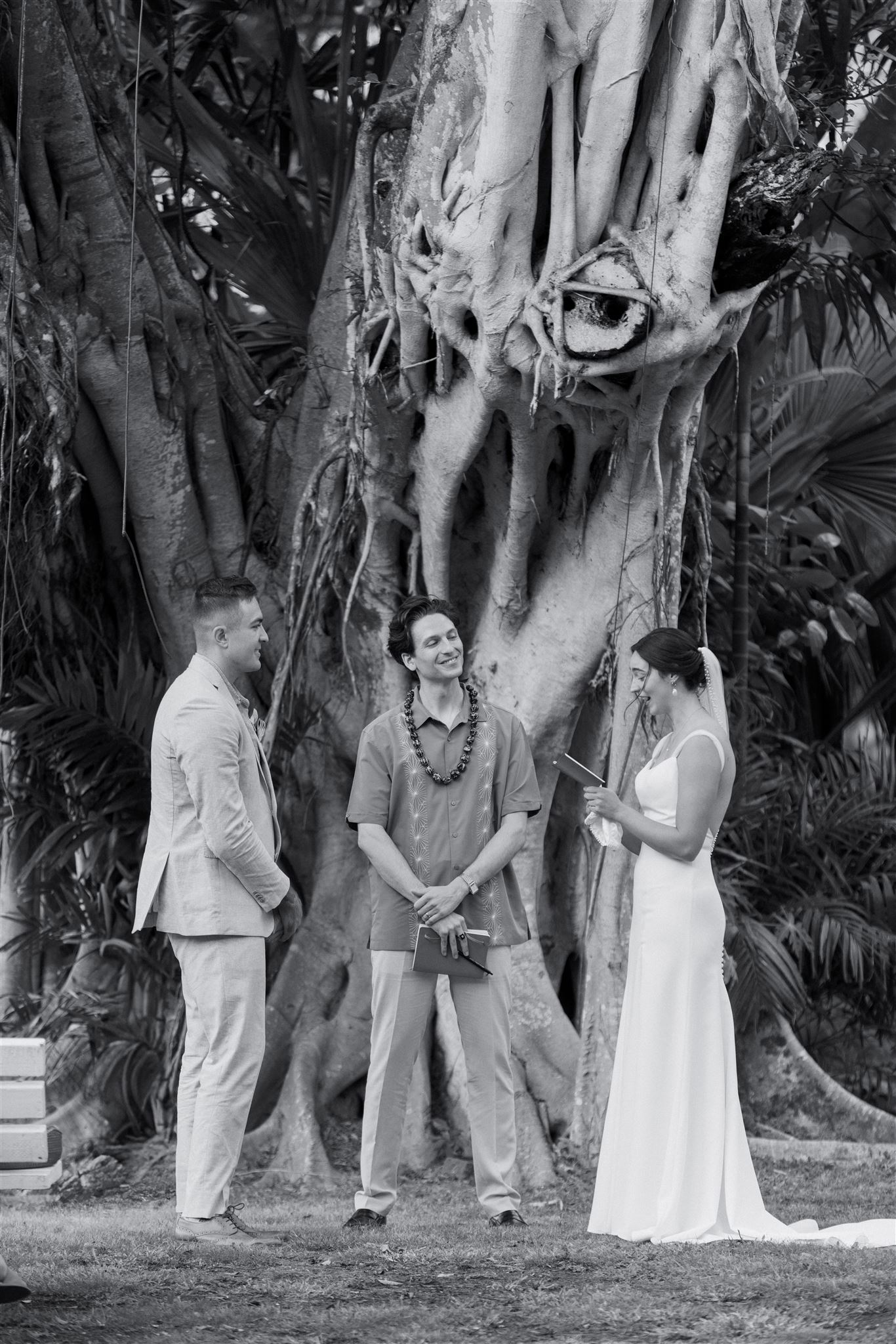 a bride and groom reading vows to each other in front of a large tree next to their officiant