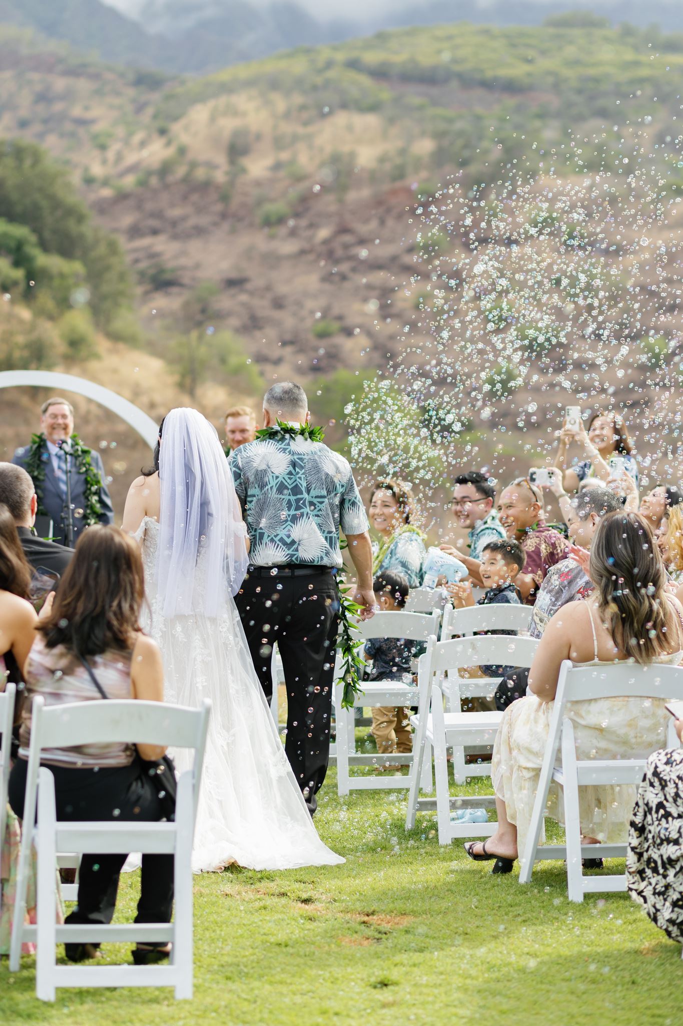 a bride walking with her father arm in arm down an aisle to her wedding ceremony spot as their guests watch them and blow bubbles