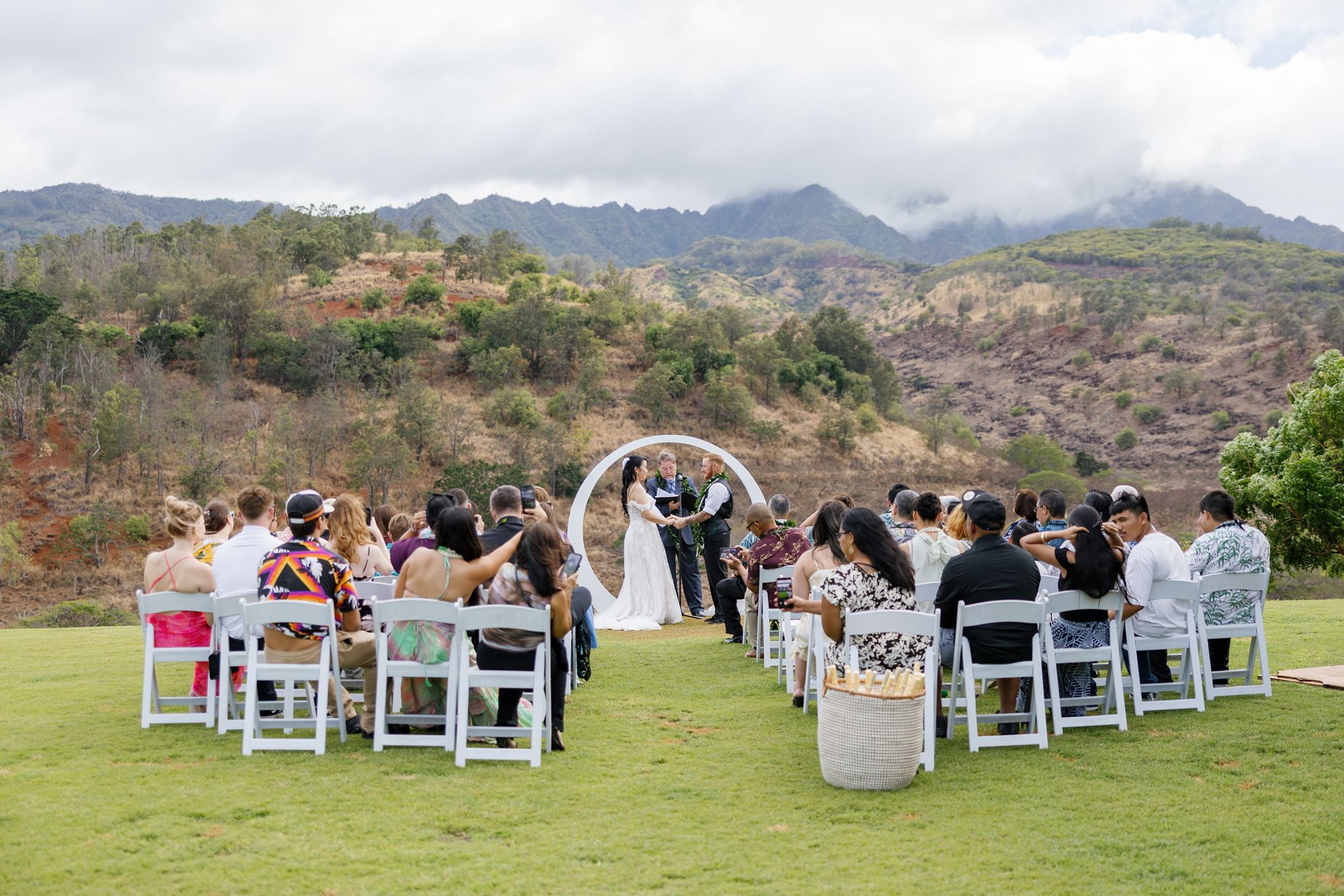 a bride and groom holding hands at their wedding ceremony as their guests watch and are seated in front of them they are surrounded by mountains