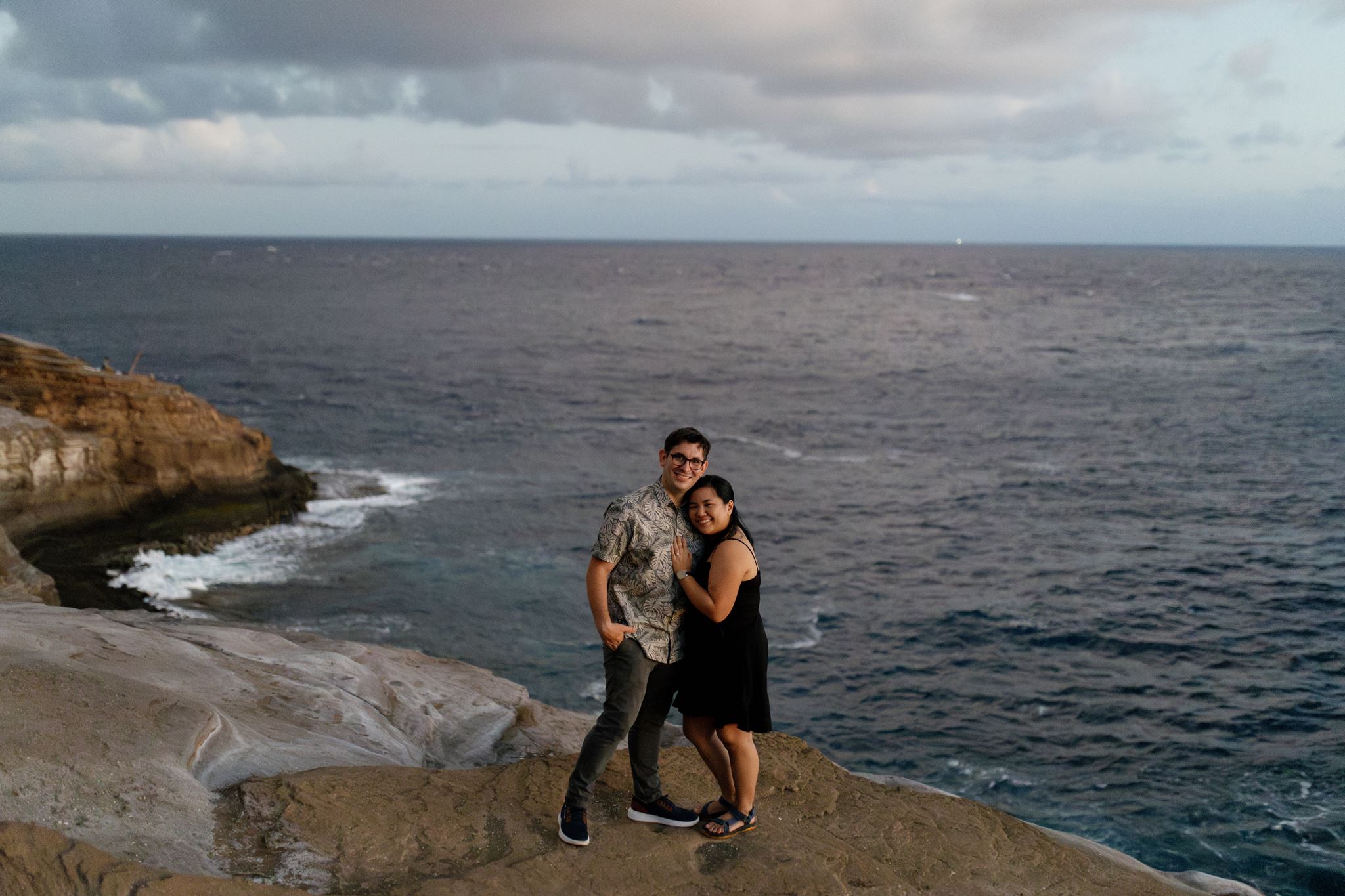 a man and woman on a cliffside next to the ocean at sunset for an engagement session