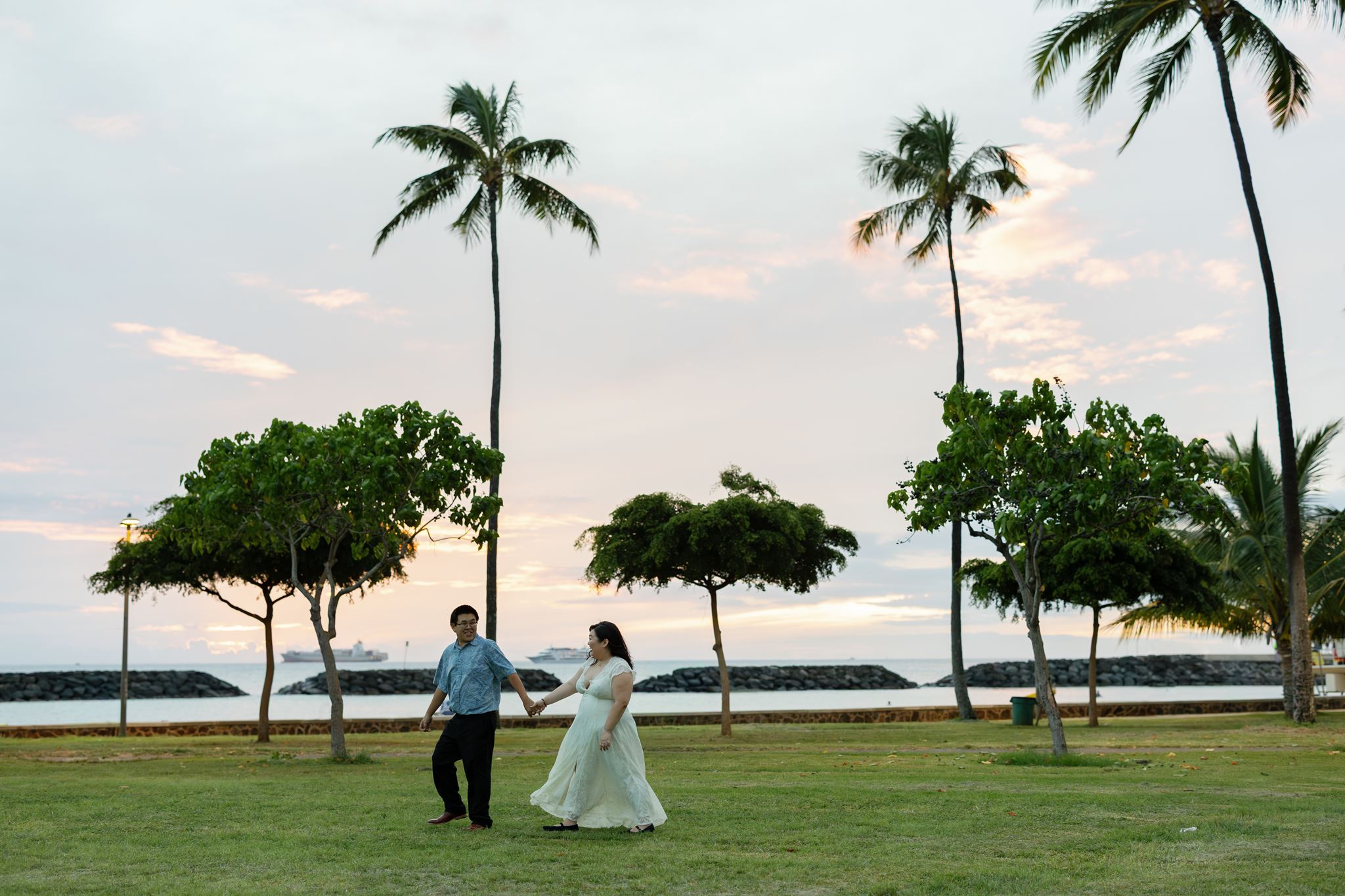 a man and woman walking across a lawn on magic island next to the water during their engagement session