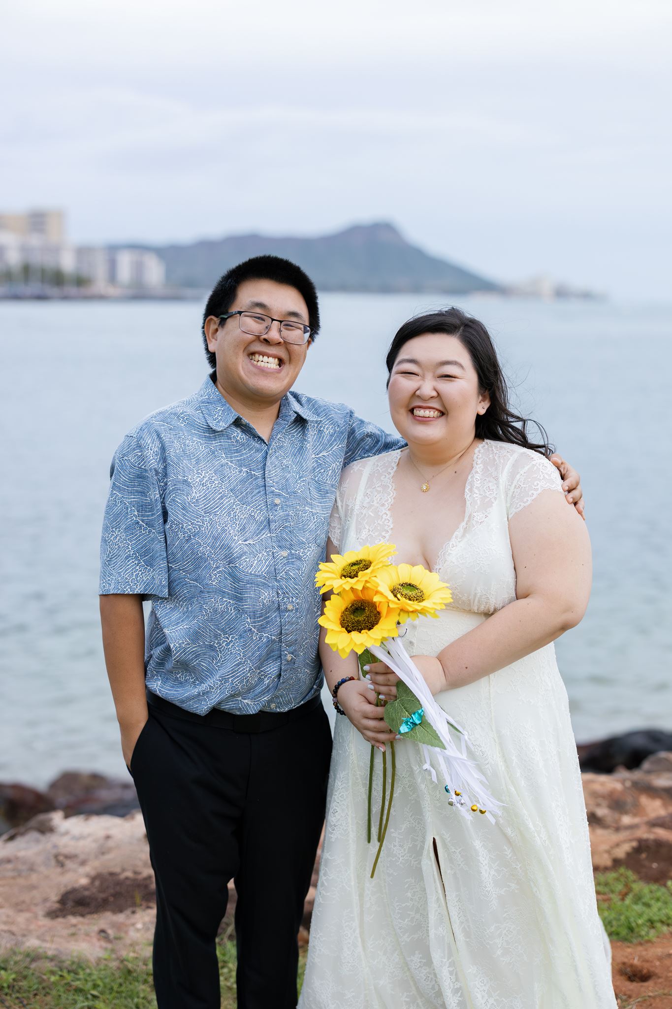 a man with his arm around his partner they are both laughing the woman is wearing a white dress and holding three sunflowers