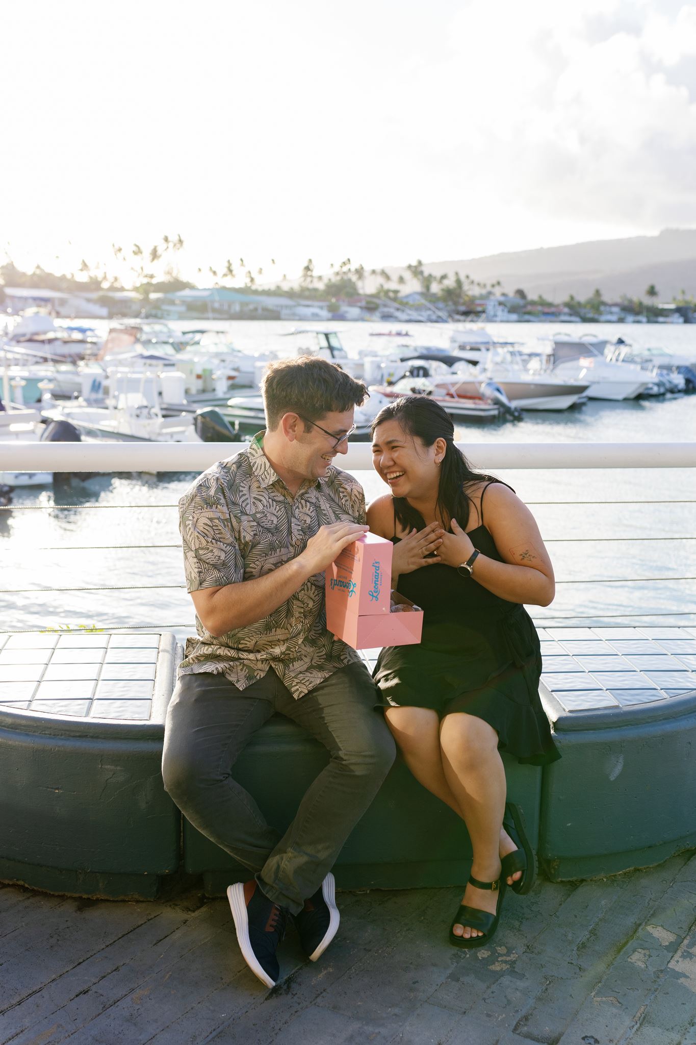 a man and woman sitting on a bench in front of a boat dock the man is holding a pink box and the woman is laughing
