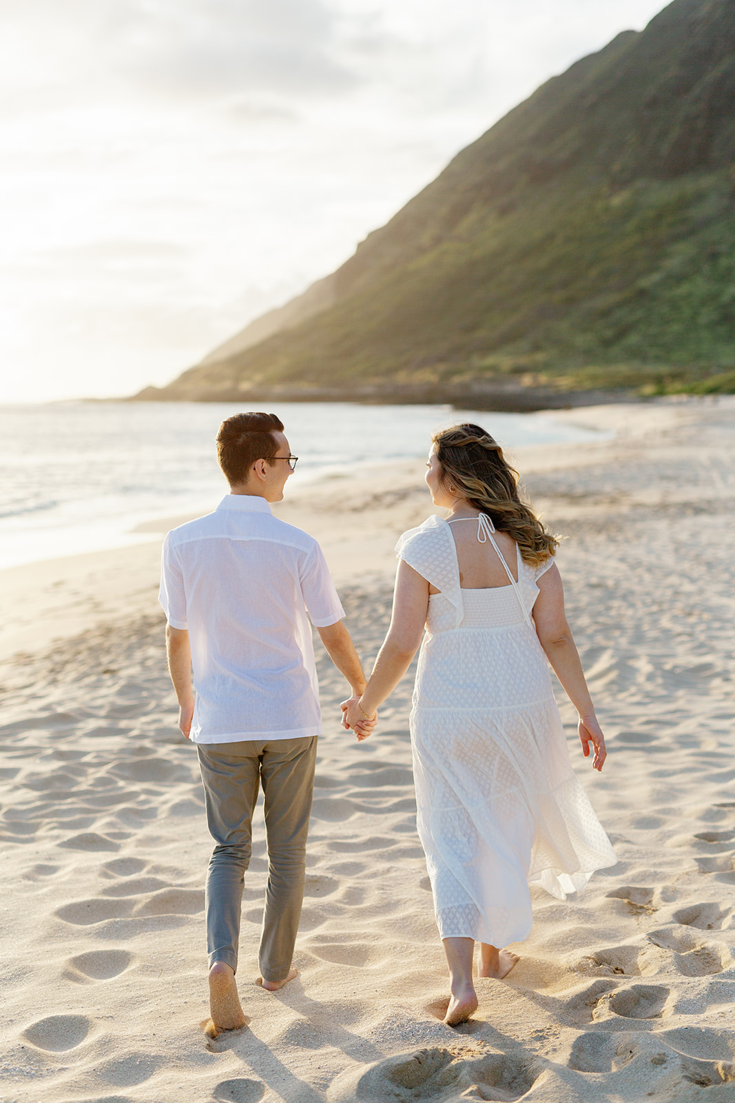 a couple having an engagement session on the beach in hawaii at sunset they are holding hands and walking down the beach together looking at one another