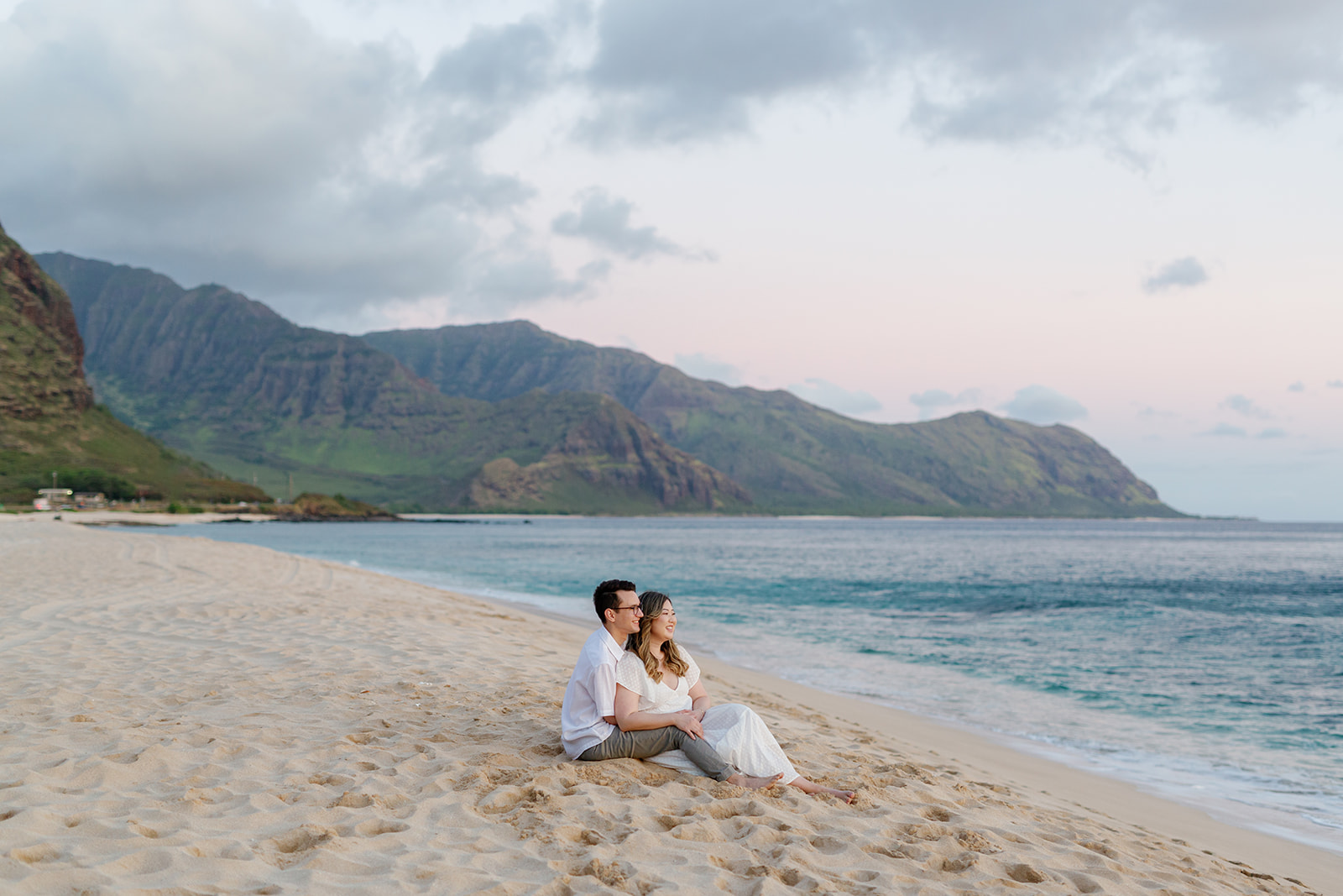 a couple sitting together in the sand during their engagement session in hawaii watching the sunset and smiling